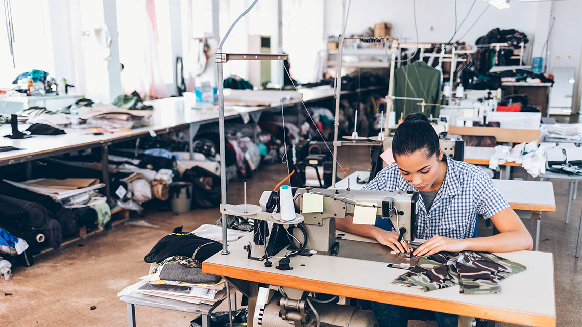 There is an adult sitting at a table with a sewing machine.  They are sewing fabric to create an article of clothing.
