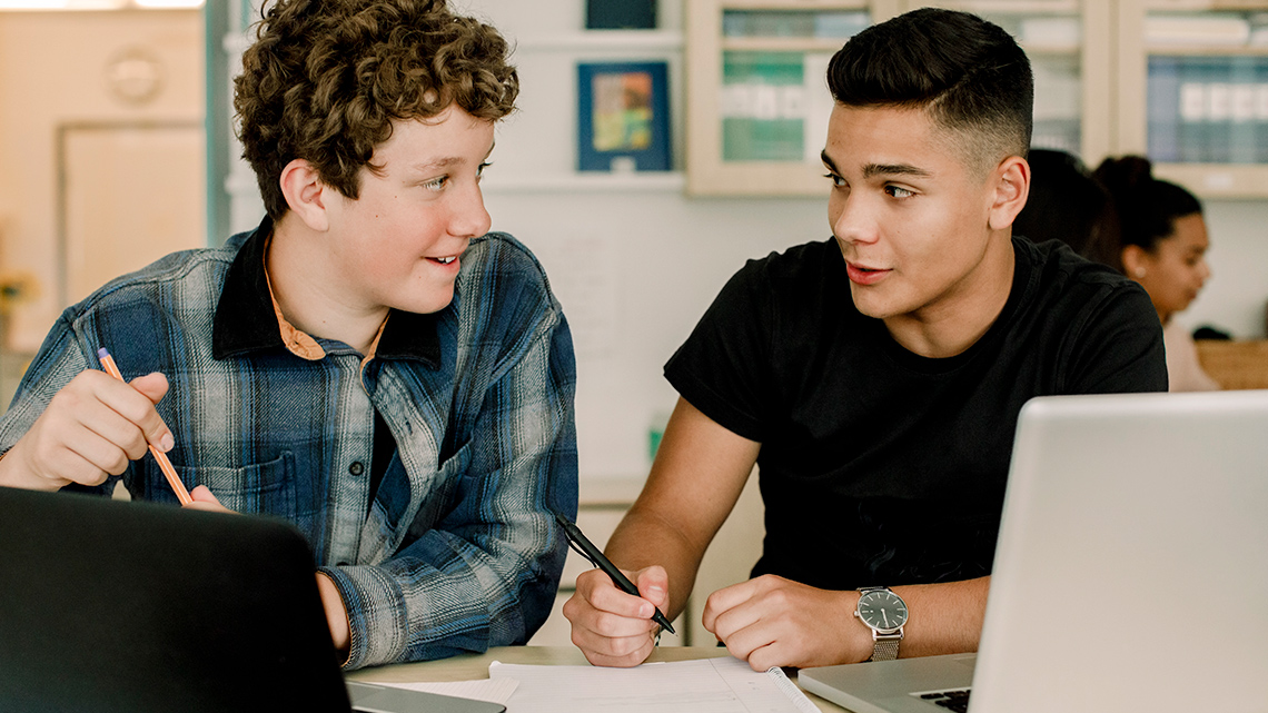 Two students working on their computers and talking to each other.