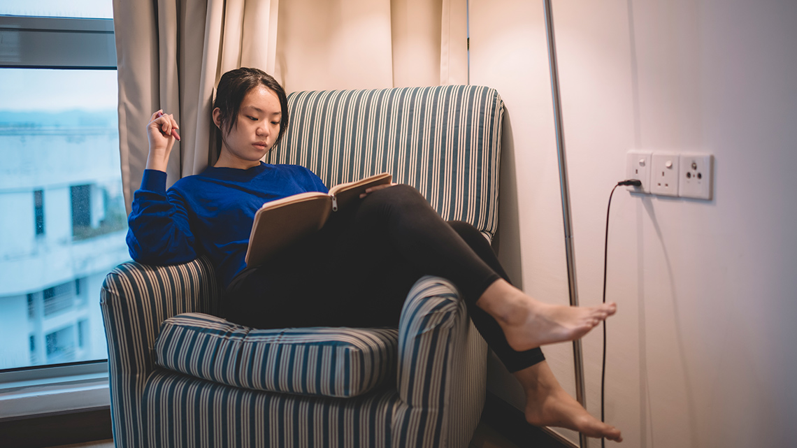 A person sitting across of a couch reading under a lamp.