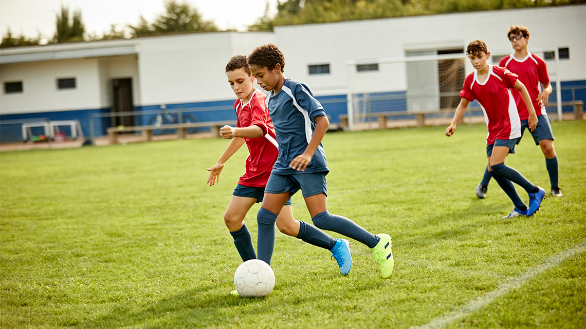 Four children running through a field after a soccer ball. One of the children is kicking the ball forward.