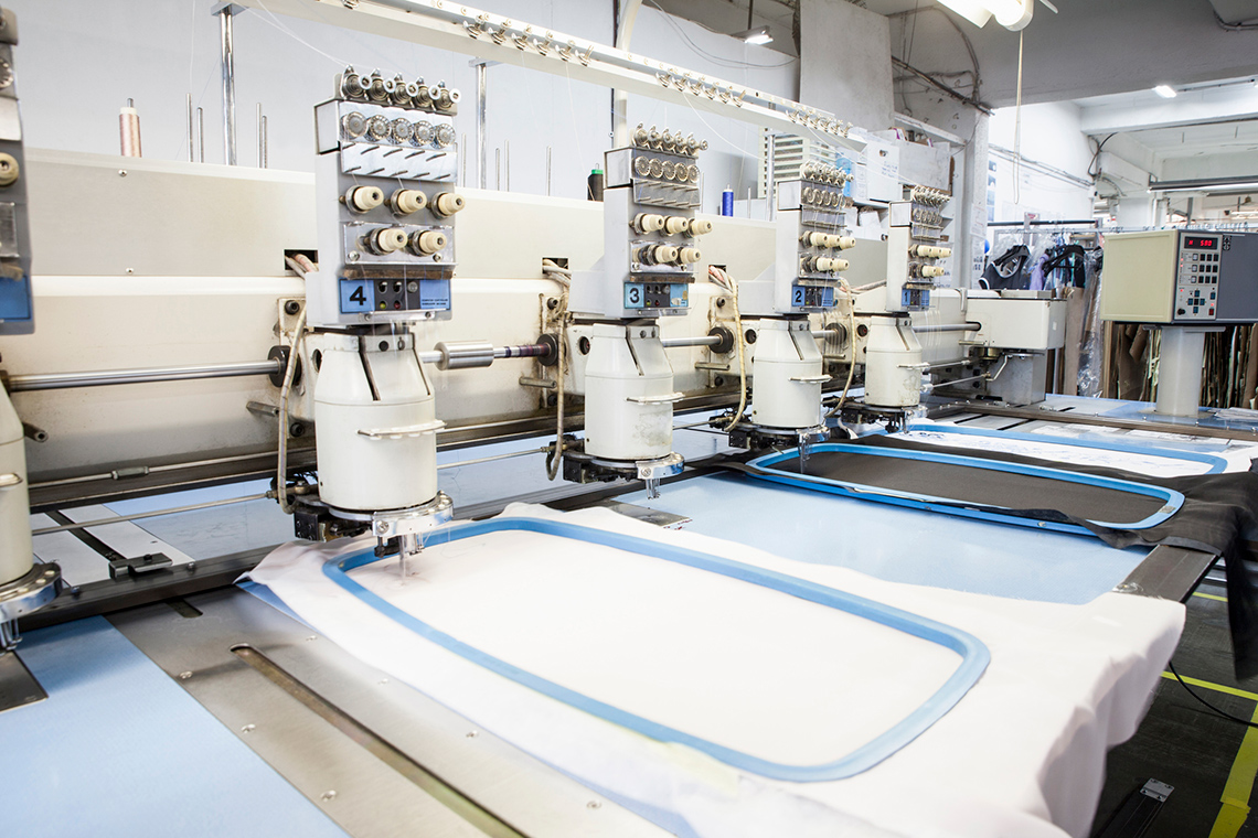 Image 2: Five automated stitching machines attached to a single beam, operating side by side. Each machine is stitching a material.
