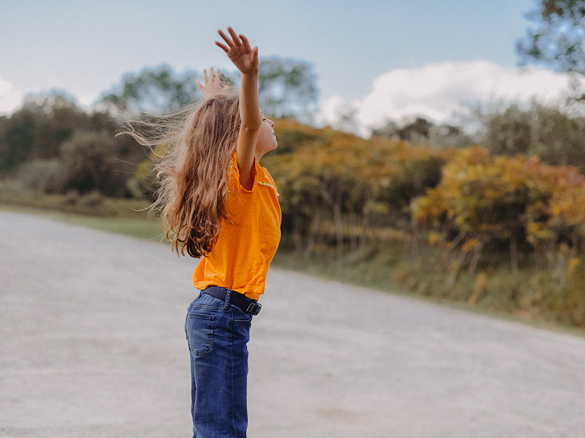 Child standing outside on a path by nature. Their arms are in the air and they are taking a deep breath.