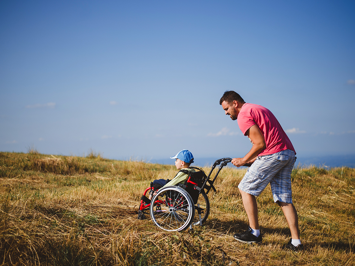 An adult is pushing a child who uses a wheelchair through a grassy field on a sunny day.