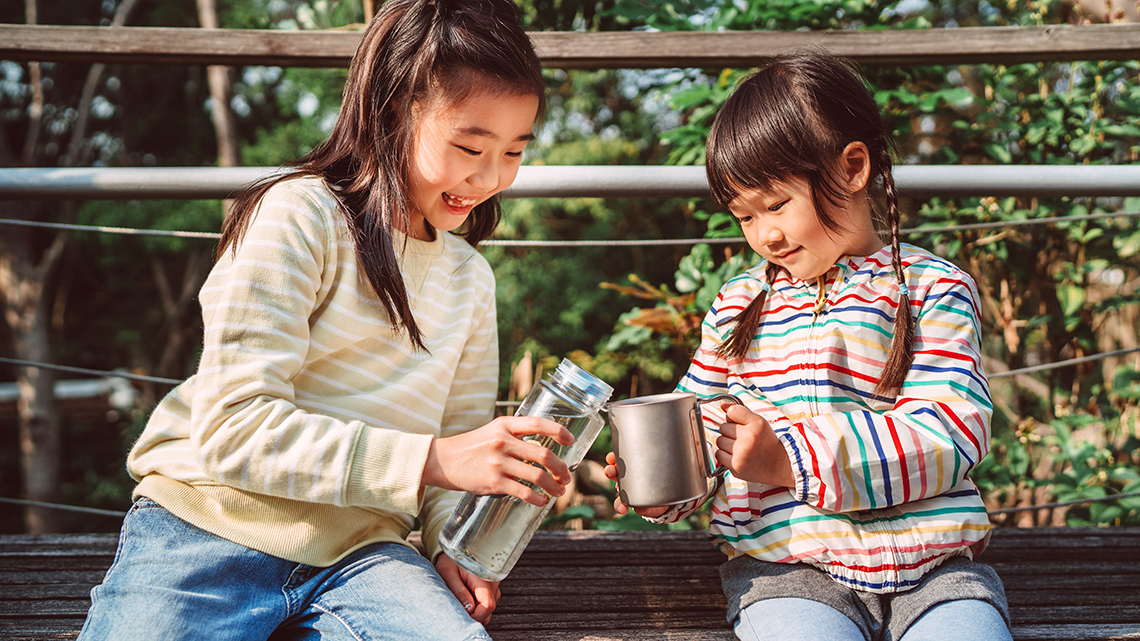 Clean Water A child is pouring clean water out of a container into a mug for another child. They are sitting together on a wooden bench outside.