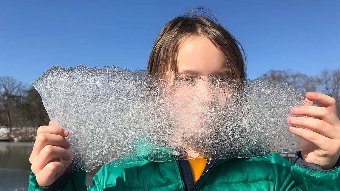 A child holding a block of ice in front of their face with their bare hands.