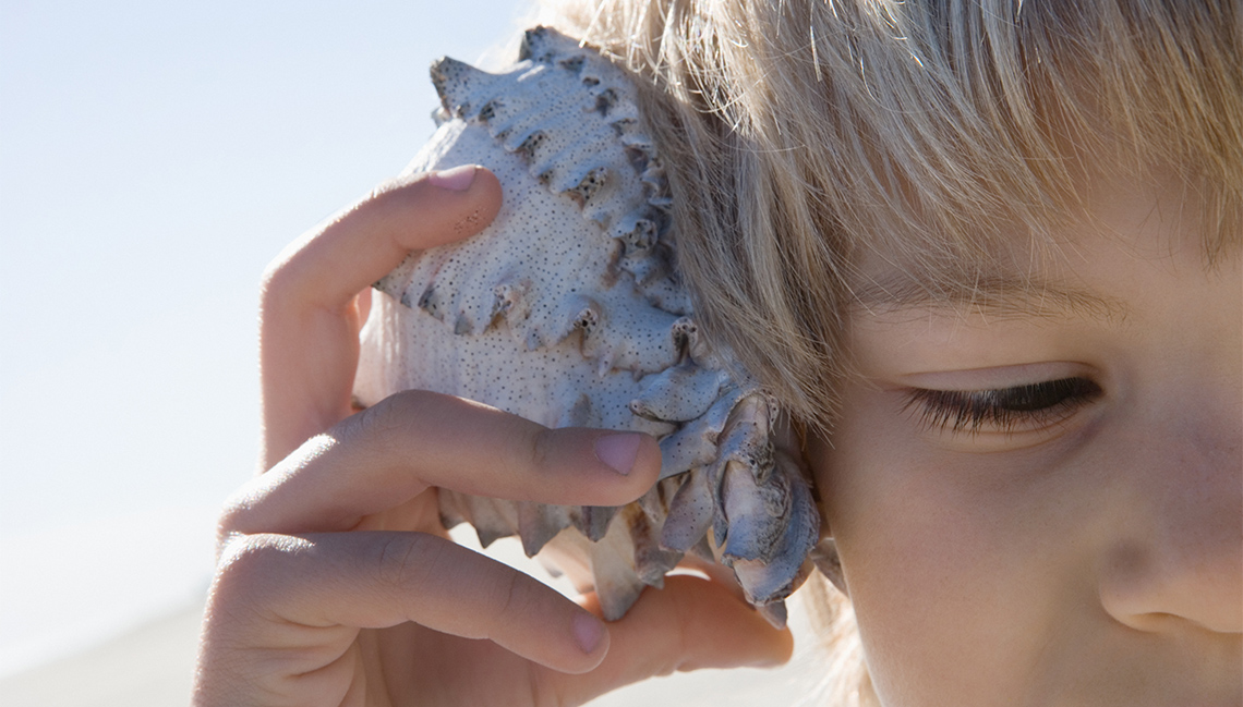 Child holds a seashell against their ear.