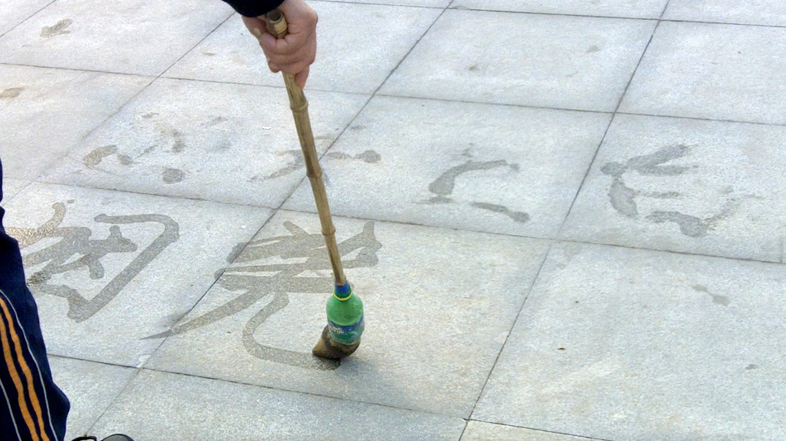 Three lines of Chinese water calligraphy being painted on the ground. The first two lines are disappearing.