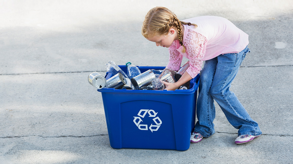 A learner is dumping cans and plastic bottles into a blue recycling container.