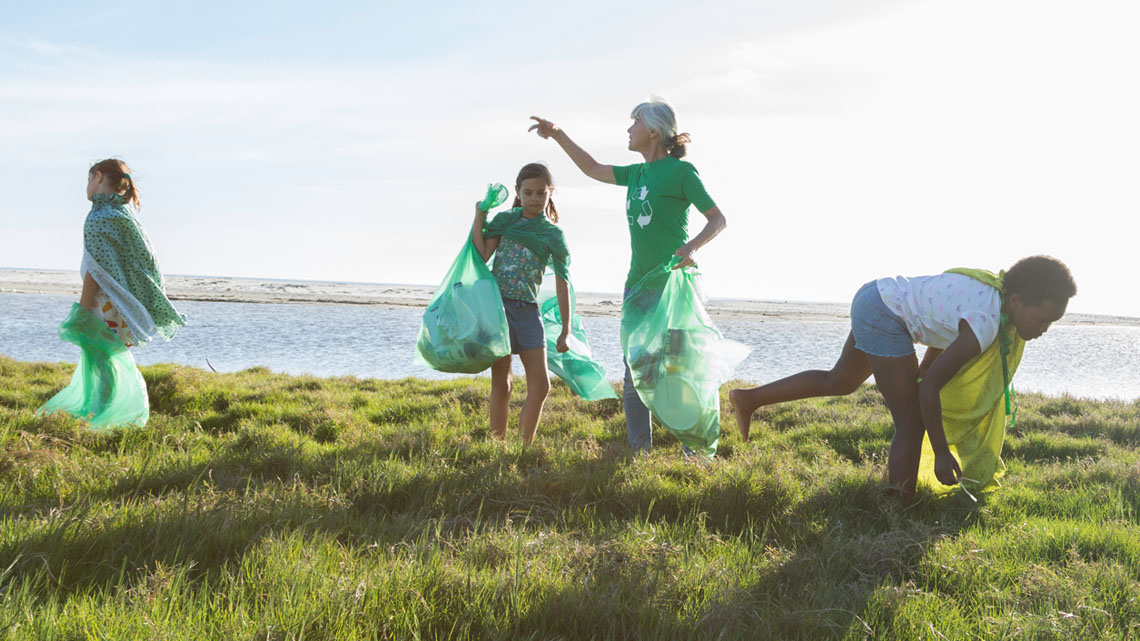 A group of young people have garbage bags and are cleaning a green space area of litter. 