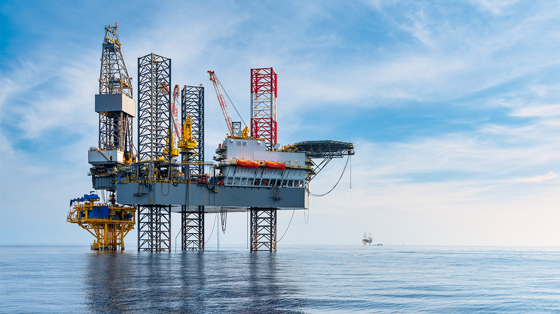 A large oil rig and drilling machinery stand in the middle of the ocean in front of a blue sky.