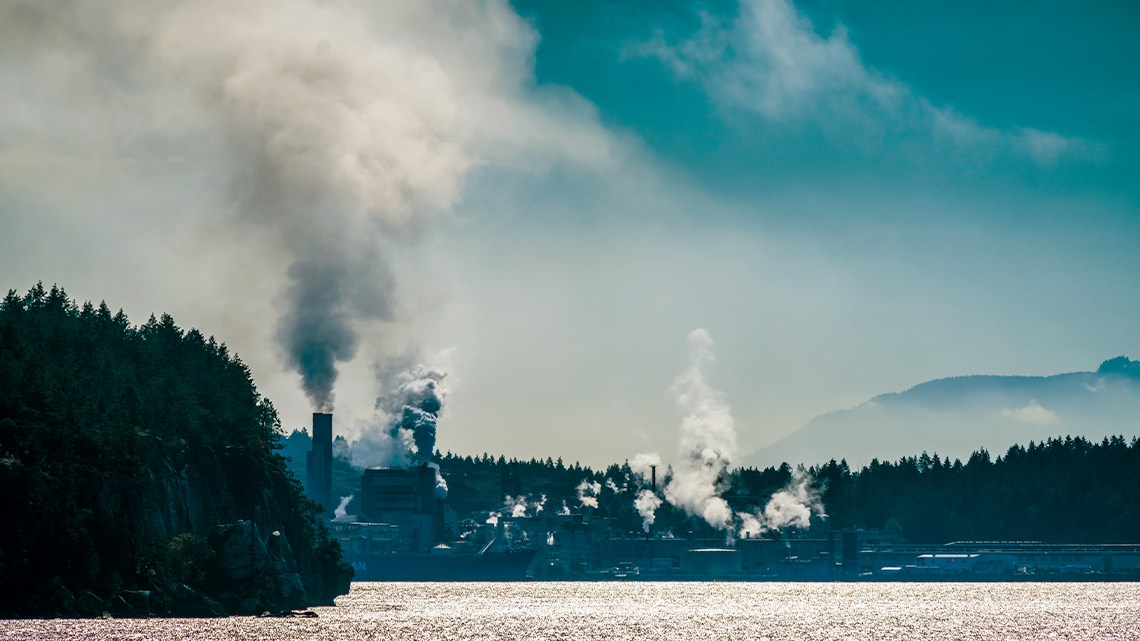 An industrial plant with smoke blowing into the air from a series of smoke stacks.