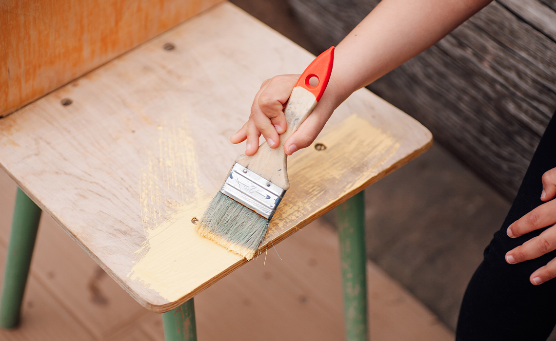 A close up of a child’s hand holding a paint brush and painting a wooden chair.