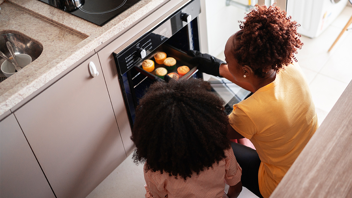 An adult and child standing by the oven removing a tray of baked goods.