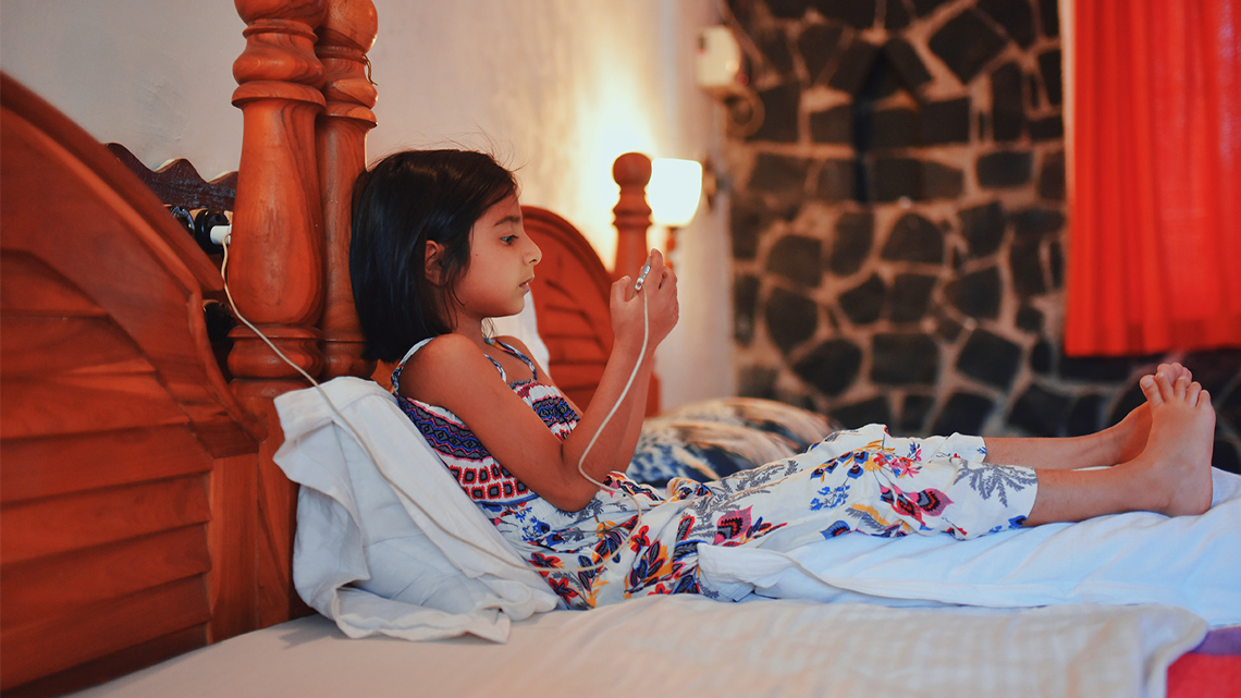 There is a child sitting on a bed with their phone plugged into the wall. There is a wall light on beside the bed.