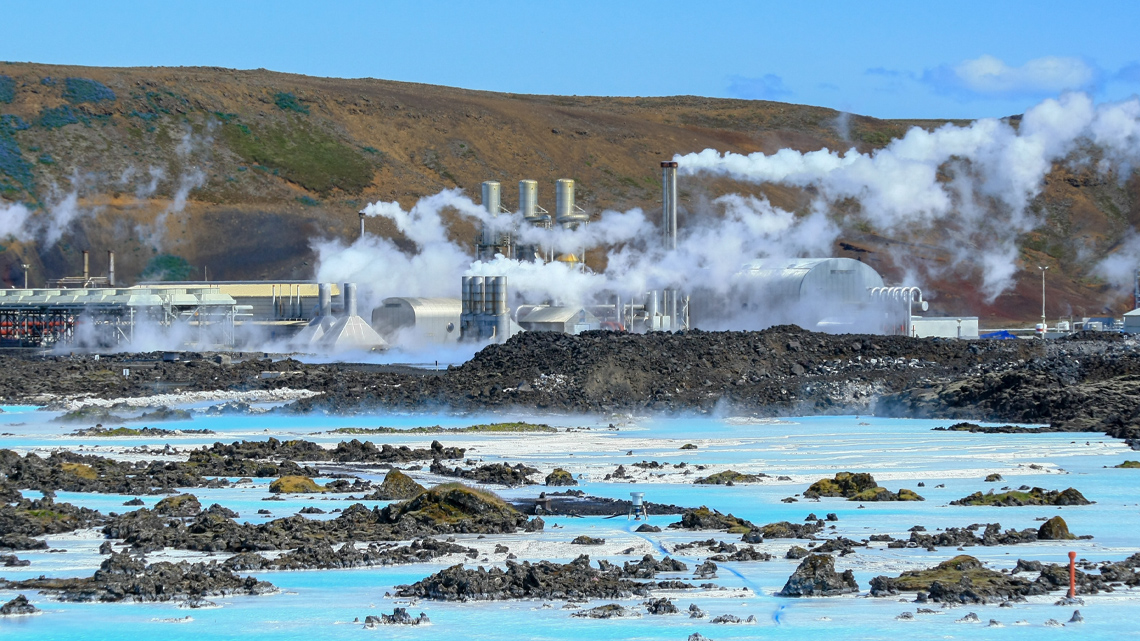 There are some buildings and long metal tubes surrounding them with steam escaping from the tops of them. There is lots of water, dirt and some hills in the background.