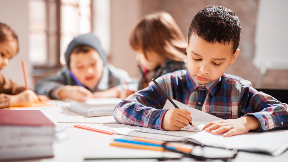 children sitting at a large desk writing in notebooks.