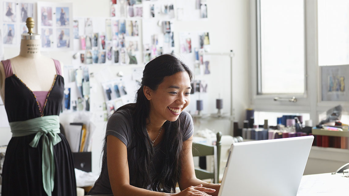 There is a young adult at their computer with a dress in the background on a mannequin. 