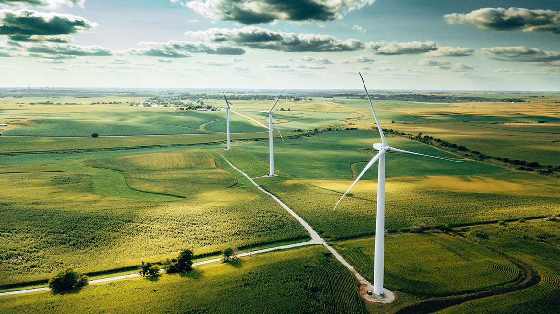 A row of three large wind turbines is pictured from the sky in a field next to two intersecting roadways. 