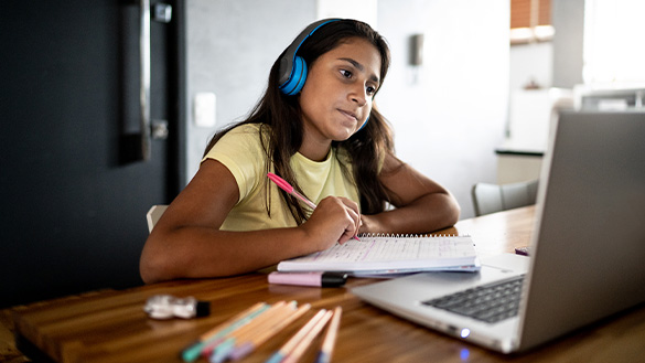 A student wearing headphones and facing their computer while writing in their notebook.