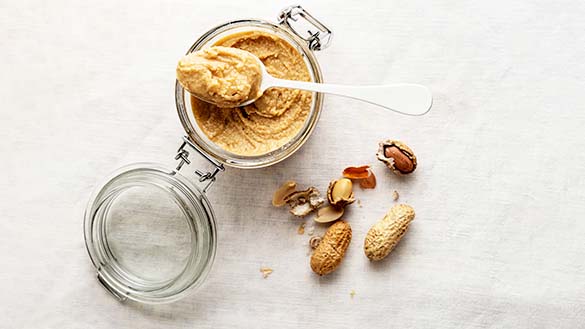 A pile of peanuts around a jar of thick brown liquid with a spoonful at the top of the jar.