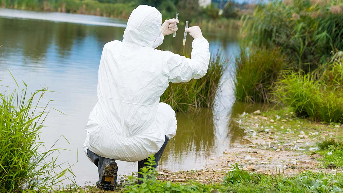 A person wearing PPE squatting on the shoreline and testing water.