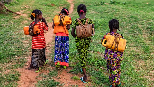 Four children walking on a dirt path while carrying water jugs.The jugs are tied to their backs with rope.