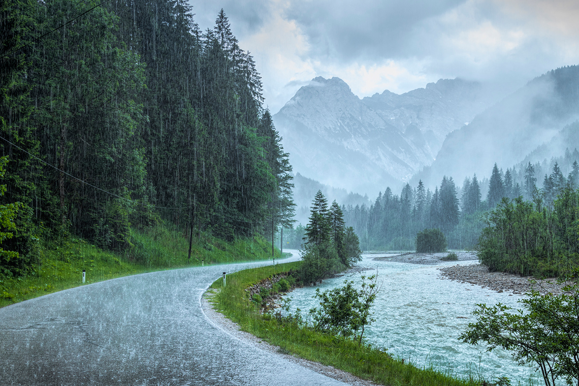 A winding road between a forest and a river, with mountains in the distance. It is raining.