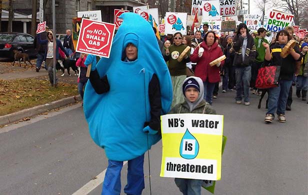 An adult dressed as a water molecule hold a sign in one hand and a child's hand in the other. A large group of protesters is behind them.