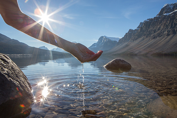 A person’s arm and hand seen scooping sparkling, clear, fresh water out of a lake beside a mountain.