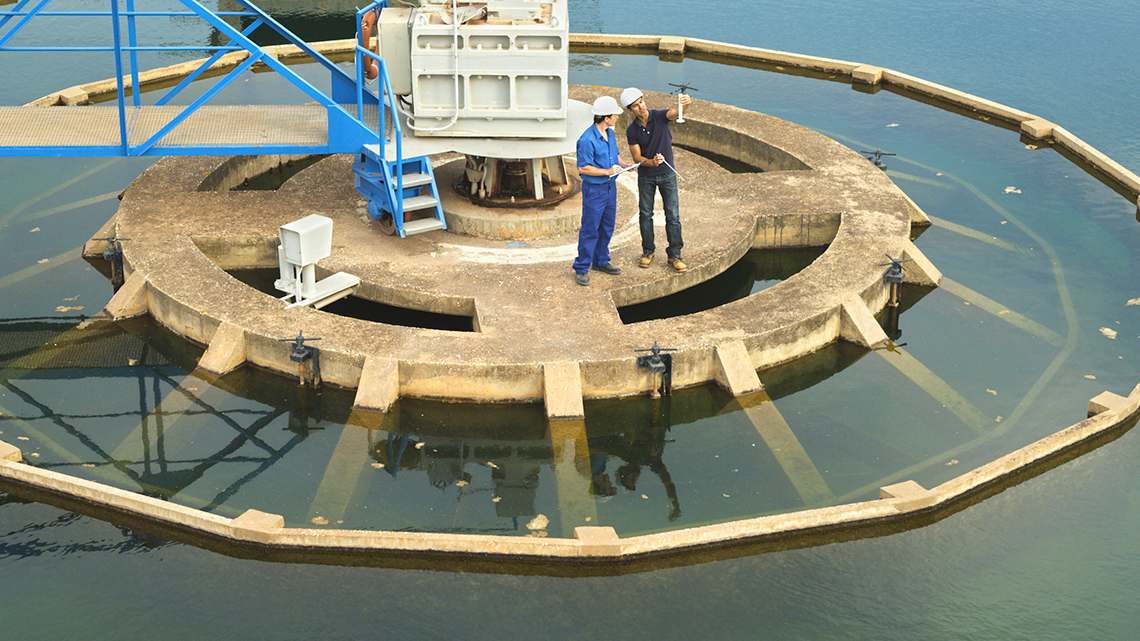 Two people wearing hard hats are standing on a concrete platform in water. They are testing water quality and recording the results.