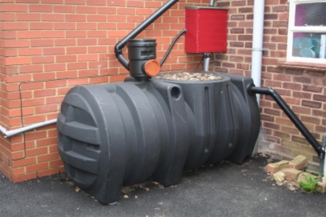 A large cylindrical tank sits beside a house. Pipes gather water from the roof.