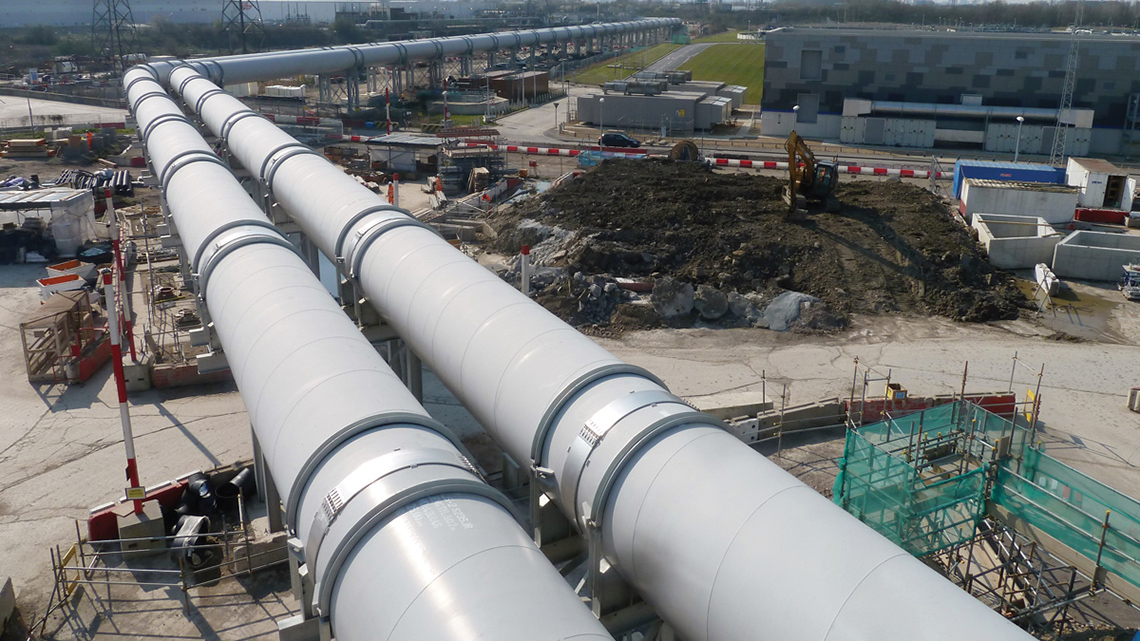 Two enormous elevated pipelines pass over a construction site and alongside city buildings.