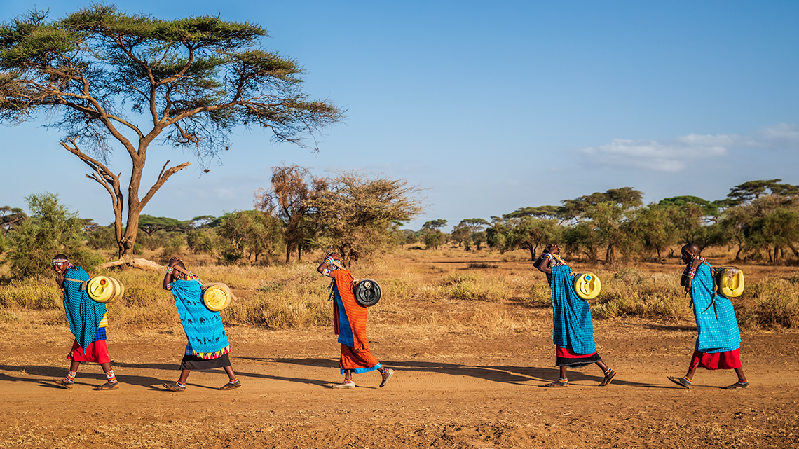 Four people walking while holding water jugs on their backs. The jugs are supported by a strap on their forehead.