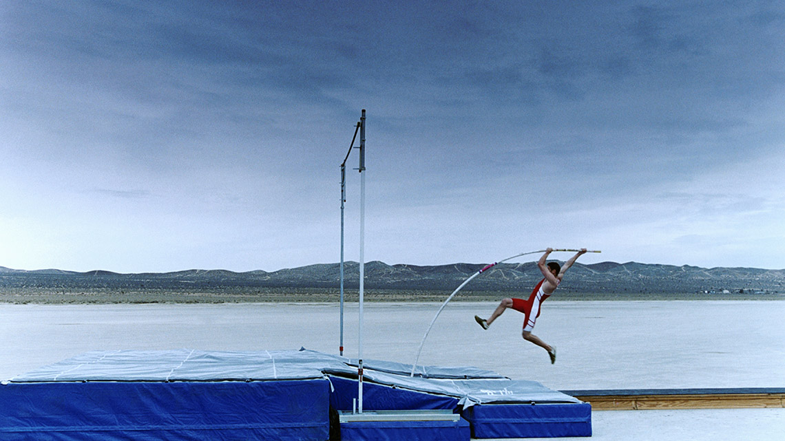 A person uses a bending vaulting pole to help push them up and over a horizontal bar.