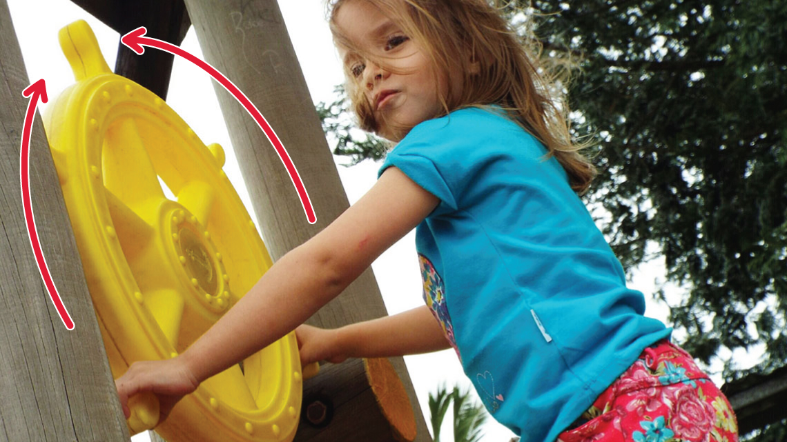 A child turns a large steering wheel that is part of a playground structure. Two curved arrows show that the wheel moves left and right. 