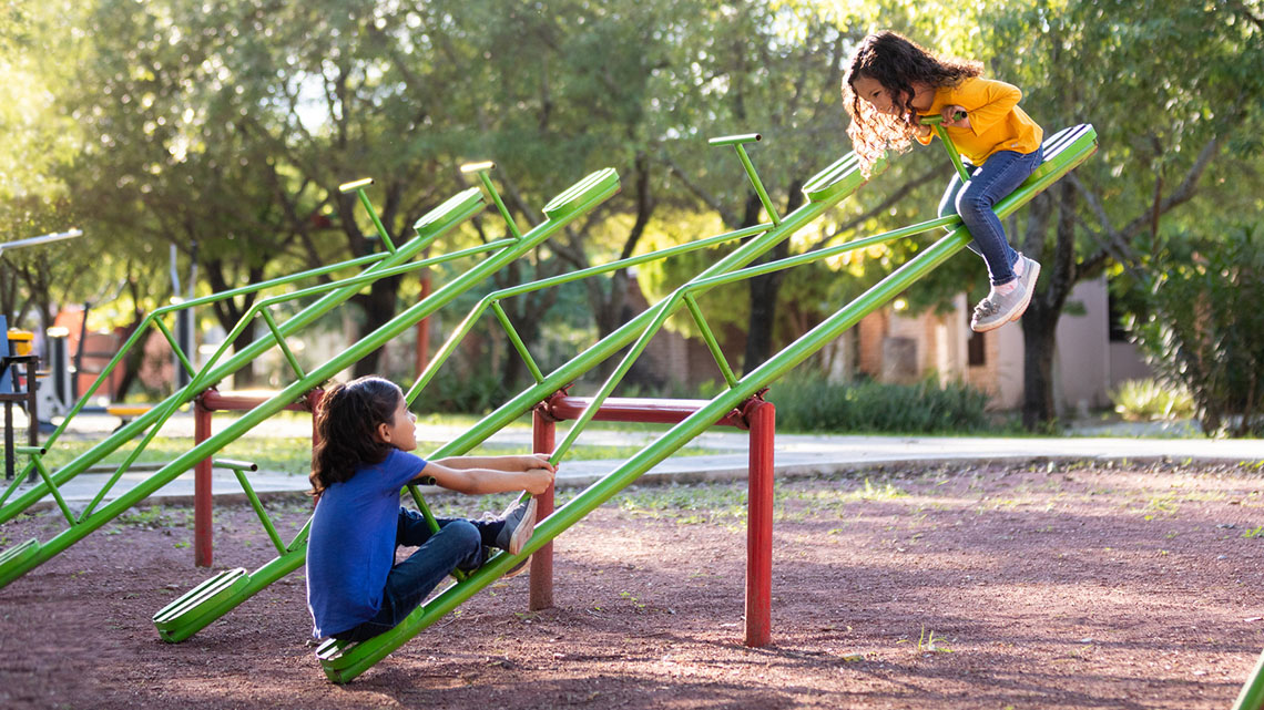 Two children are seated on opposite ends of a teeter-totter. One end is on the ground and the other end is in the air. 