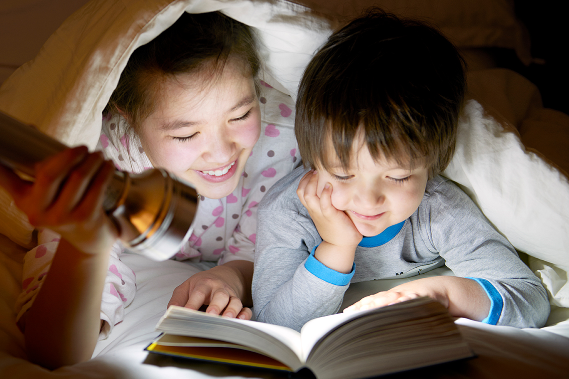 Two children with their heads covered by a blanket, reading a book together using the light of a flashlight.