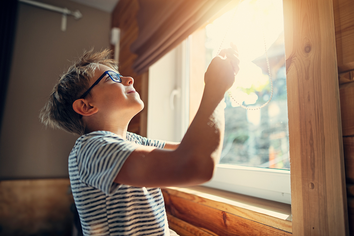 Child pulling a string to open blinds on a window to let the sunlight into a room.