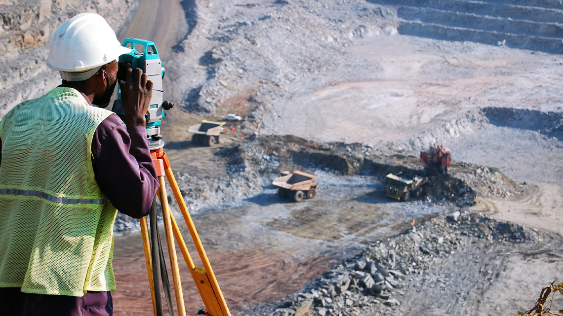 Worker standing at the top of an open pit mine looking through a surveying tool on a tripod stand.
