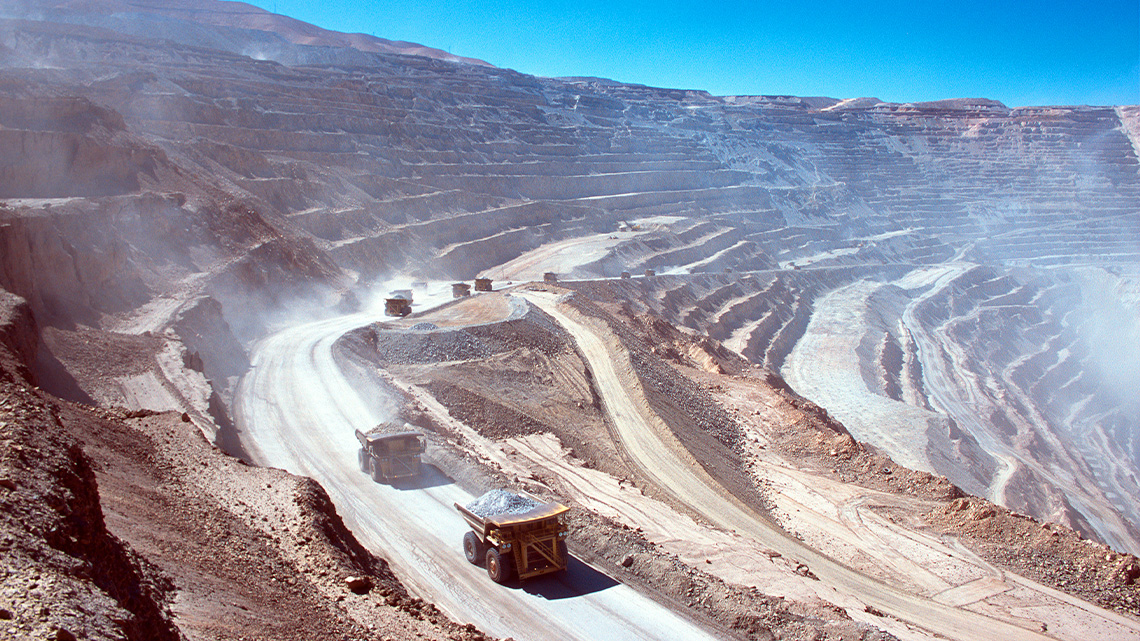 Trucks travelling down dirt roads on the side of a dusty mountain leading down into an open pit mine.
