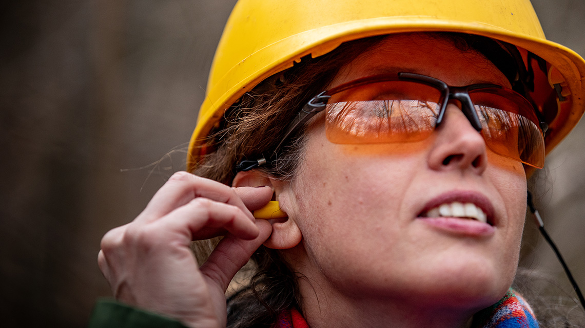 A worker in a hard hat and safety glasses is placing an ear plug into their ear.