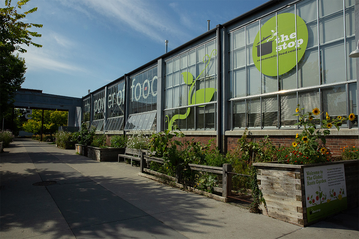 A long building with multiple glass windows. Plots with different kinds of plants are set up along the side of the buildings.