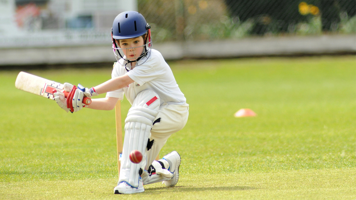 Young child in protective gear playing a sweep shot in a game of cricket. They are wearing a helmet, knee pads, shoes with cleats and a t-shirt.