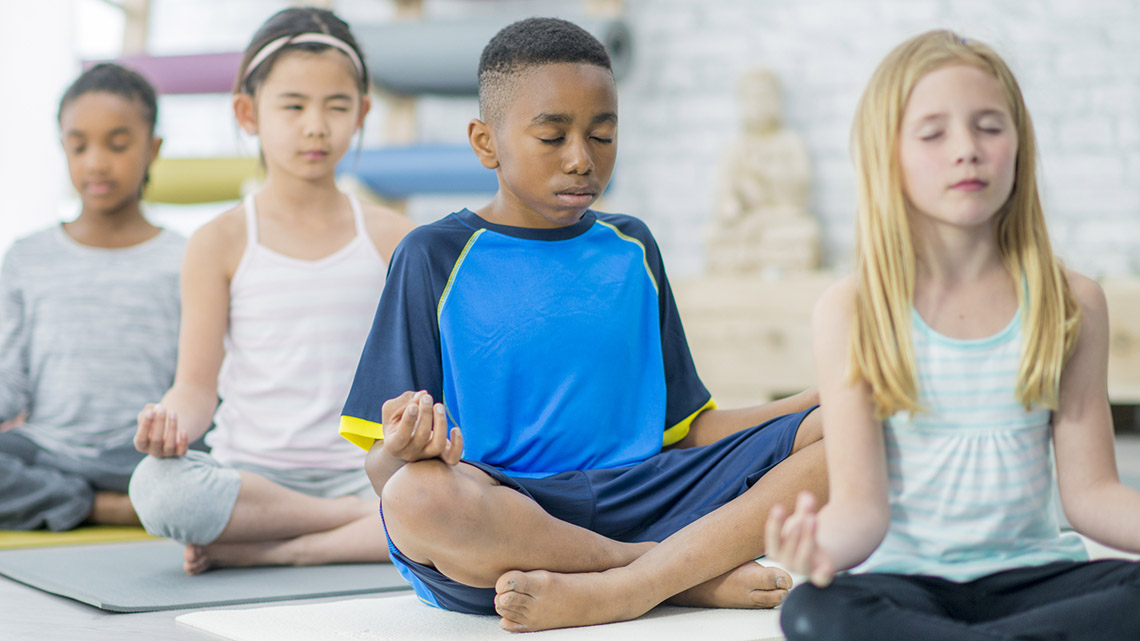 Children sitting on mats with their legs crossed, their hands on their knees, their eyes closed and breathing in and out.