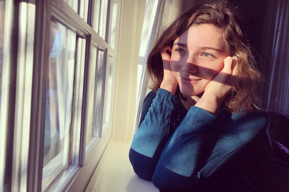 A person with their eyes open, smiling and soaking up the Sun while looking out the window in a home.