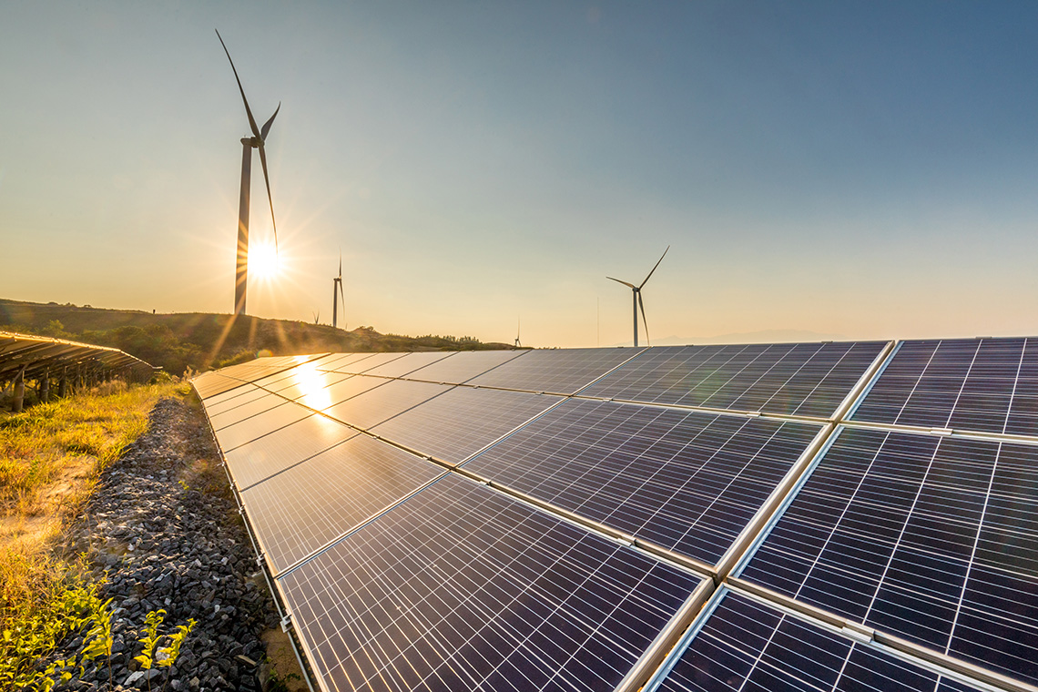 Sunset hitting a row of solar panels with wind turbines in the background.