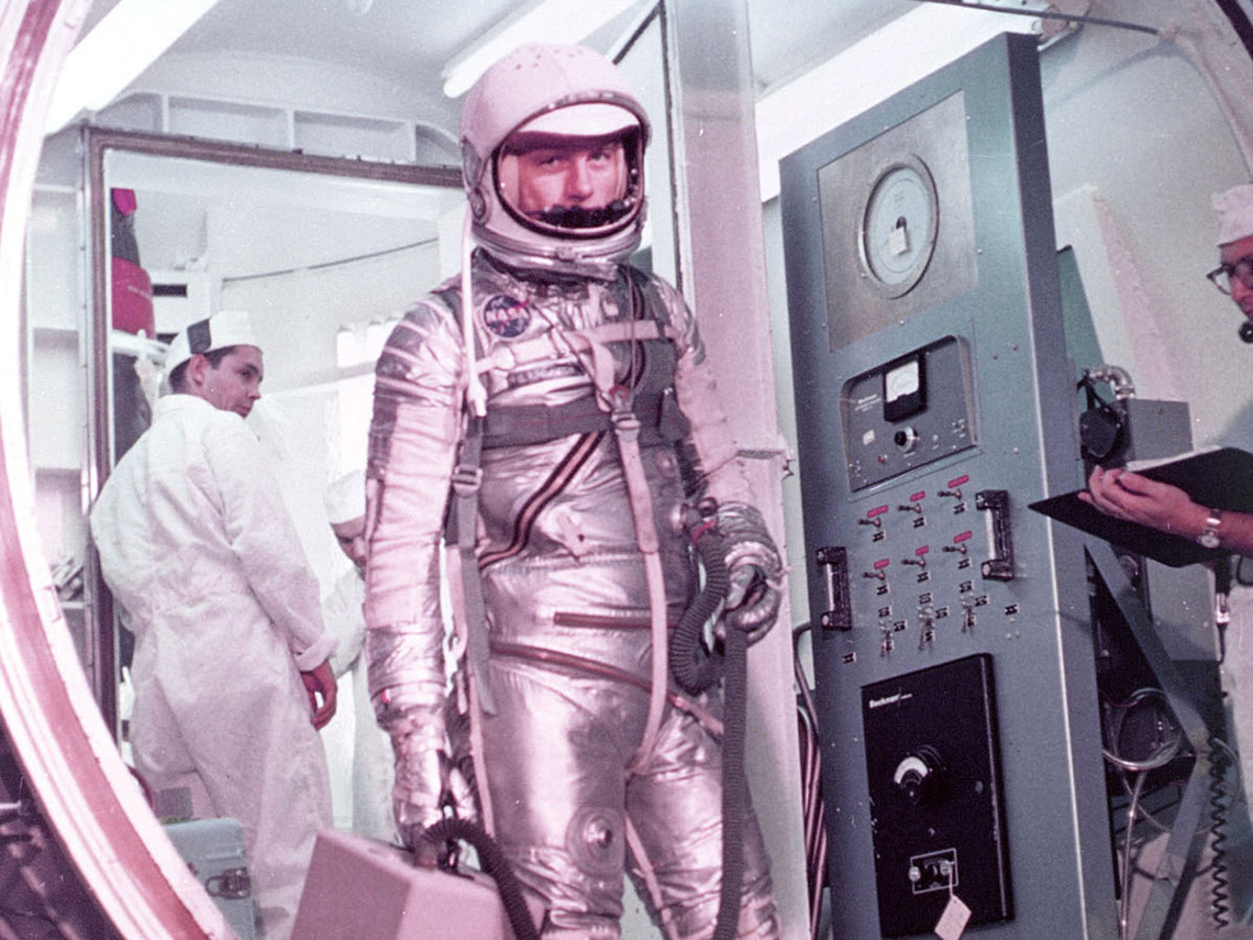 John Glenn standing at the door to a spacecraft wearing a fitted, shiny silver spacesuit.