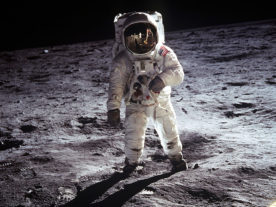 Astronaut Buzz Aldrin standing on the Moon wearing a bulky spacesuit with a large backpack and a reflective helmet.