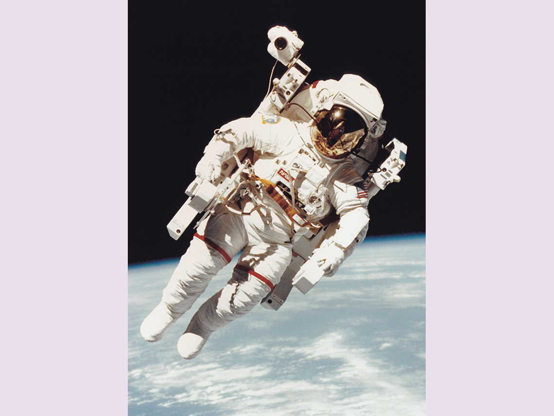 Astronaut floating in space wearing a bulky spacesuit with very large mechanical backpack device with hand controls.