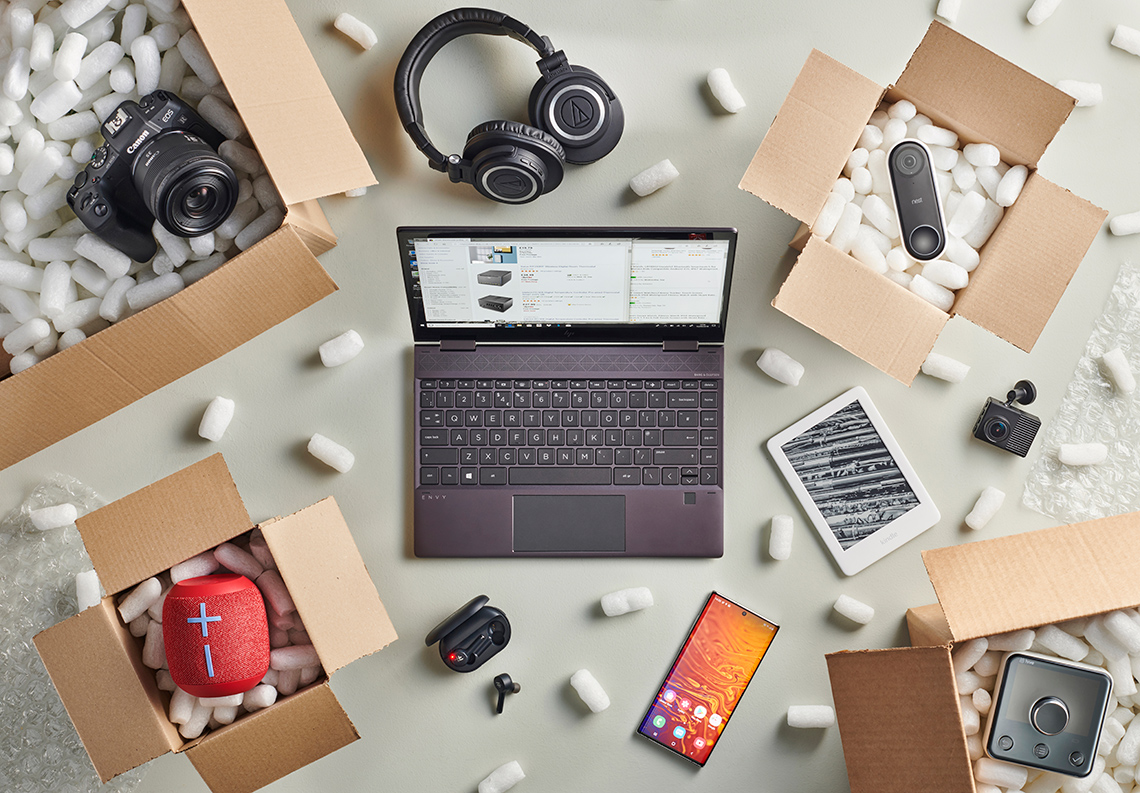 A laptop, tablet, phone, cameras, speakers, headphones, ear buds, and thermostat sitting on a table.